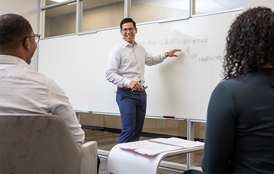 a man and a woman seated, another man smiling and pointing to whiteboard 