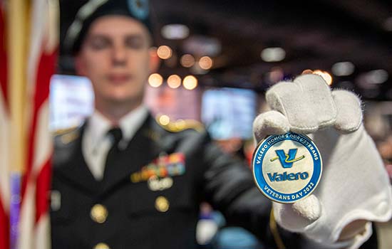In the background, a ROTC member holds an American flag in his right hand, and in the foreground, his left hand displays a Valero "challenge coin" in honor of Veterans Day 2022.