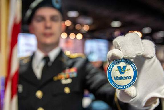In the background, a ROTC member holds an American flag in his right hand, and in the foreground, his left hand displays a Valero "challenge coin" in honor of Veterans Day 2022.