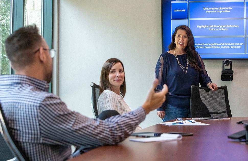 Three individuals in a conference room, one standing next to presentation screen