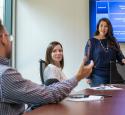 Three individuals in a conference room, one standing next to presentation screen
